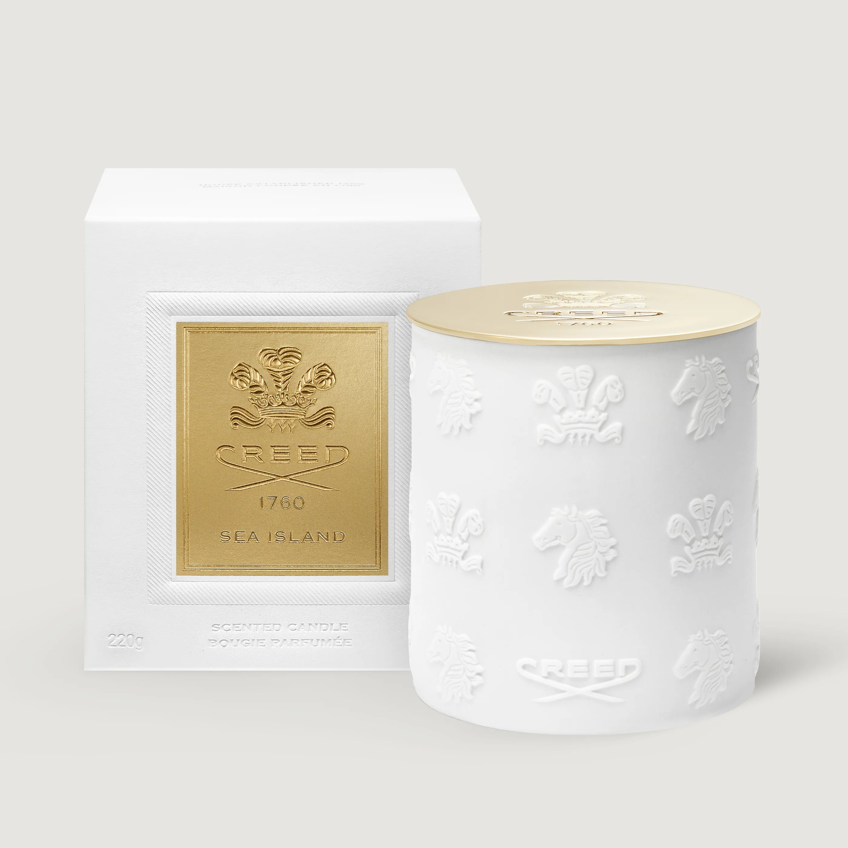 A white ceramic candle jar with embossed crest motifs and a gold lid, shown next to its white box packaging featuring a gold crest label. The brand name 'Creed' is visible on both the box and the jar.