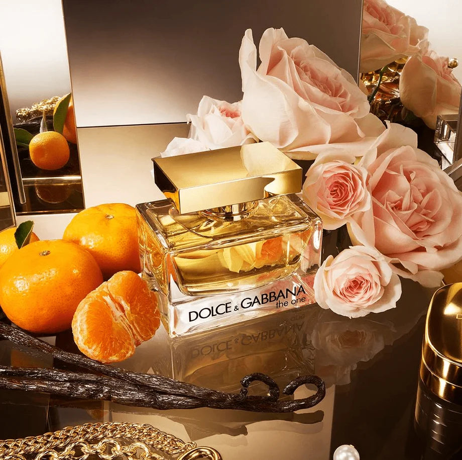 Dolce & Gabbana 'La Gioia' perfume bottle with a gold cap, styled on a reflective dark surface with pink roses, oranges, vanilla beans, and a pearl.