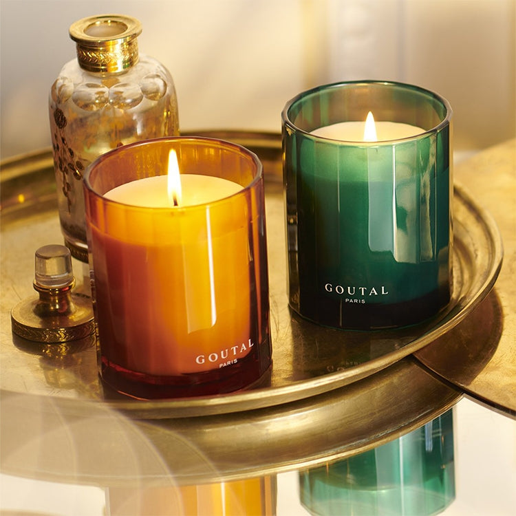 Two lit Goutal Paris scented candles in amber and emerald green glass holders, styled on a round brass tray with a decorative glass bottle in the background.