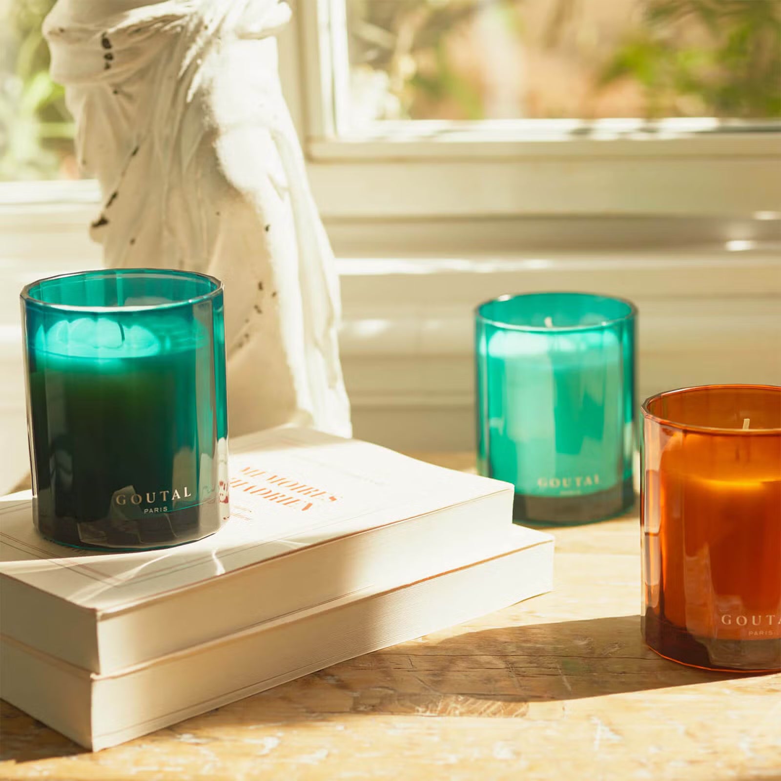 Three Goutal Paris scented candles in different colored glass jars—teal green, teal blue, and amber orange—arranged on a wooden table with stacked books in a sunlit room.