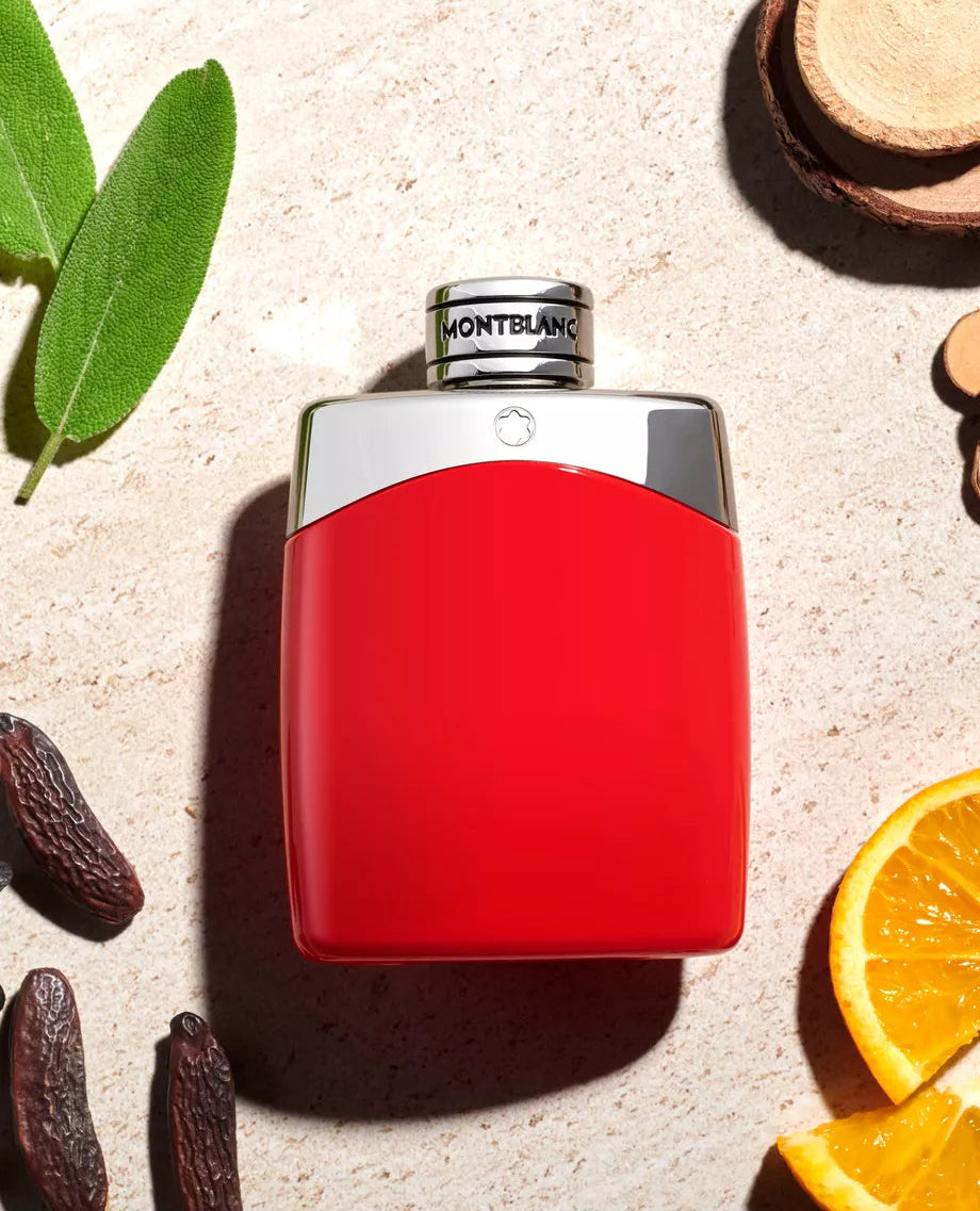 A red and silver Montblanc perfume bottle, styled on a textured beige stone surface with sage leaves, cloves, and orange slices.