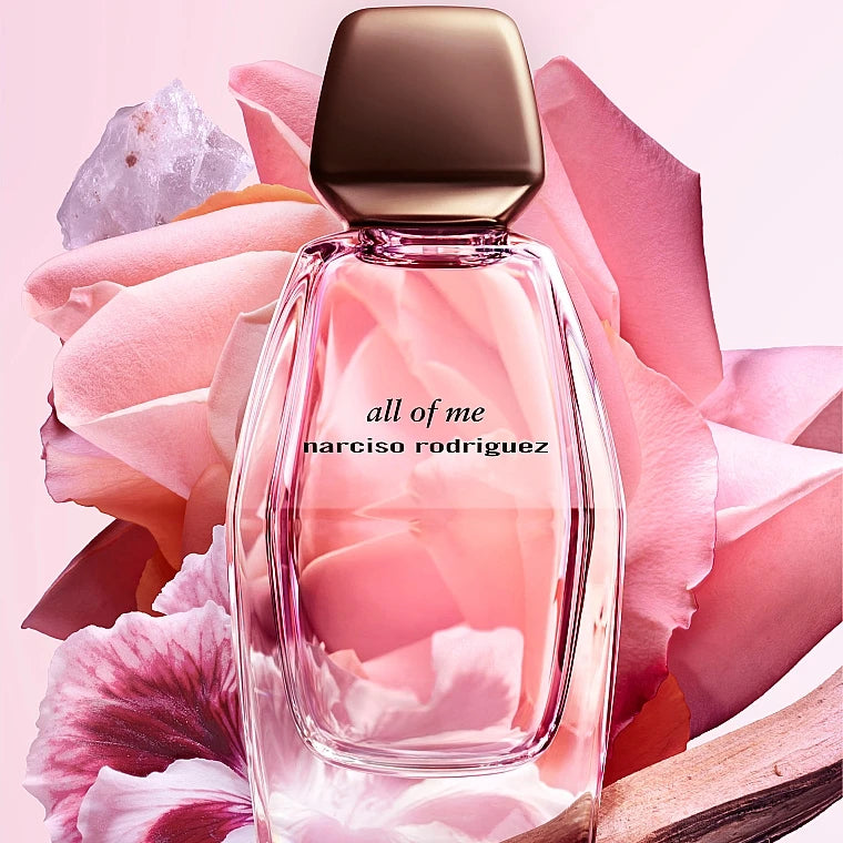 Narciso Rodriguez 'all of me' perfume bottle with a metallic bronze cap, set against a background of soft pink rose petals and a crystal.