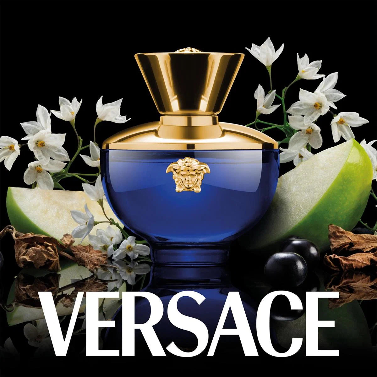 Versace perfume bottle in deep cobalt blue with a gold cap and Medusa emblem, surrounded by white flowers, green apple slices, and dark olives on a black background, with the VERSACE logo in large white text at the bottom.