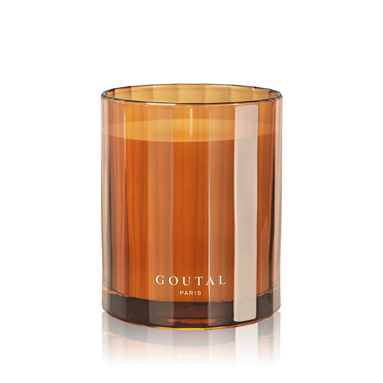 An amber glass jar candle with a white wick and the brand name 'GOUTAL PARIS' printed in white near the base, set against a clean white background with a soft reflection.