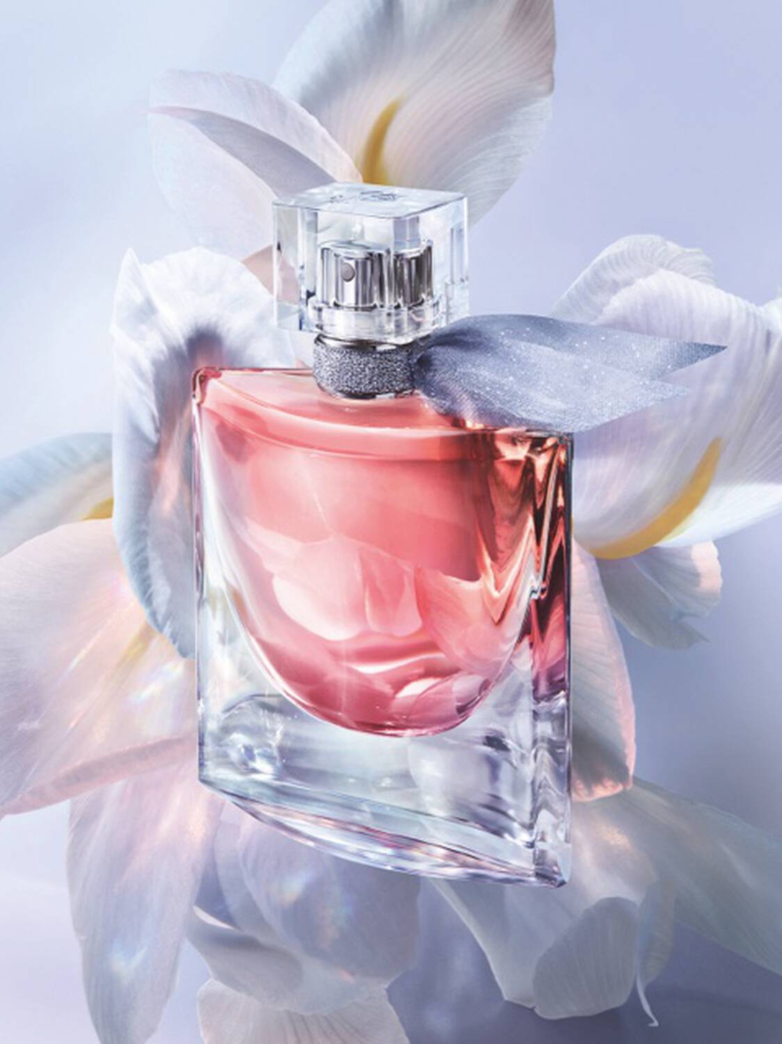 A clear glass perfume bottle filled with pink liquid, wrapped in a sheer gray ribbon, set against large white flowers on a soft lavender background.