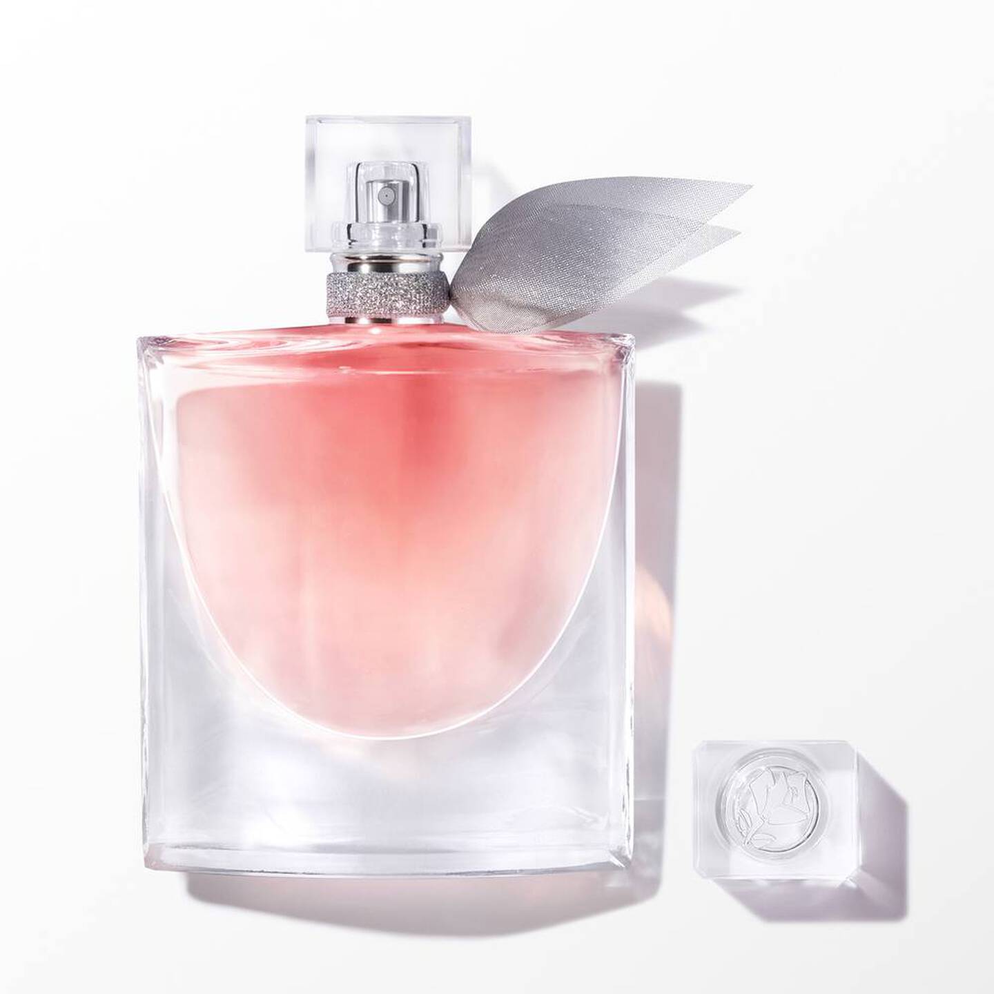 A clear glass perfume bottle with a soft pink liquid and a silver leaf-shaped cap, shown on a white background with the cap placed beside it.